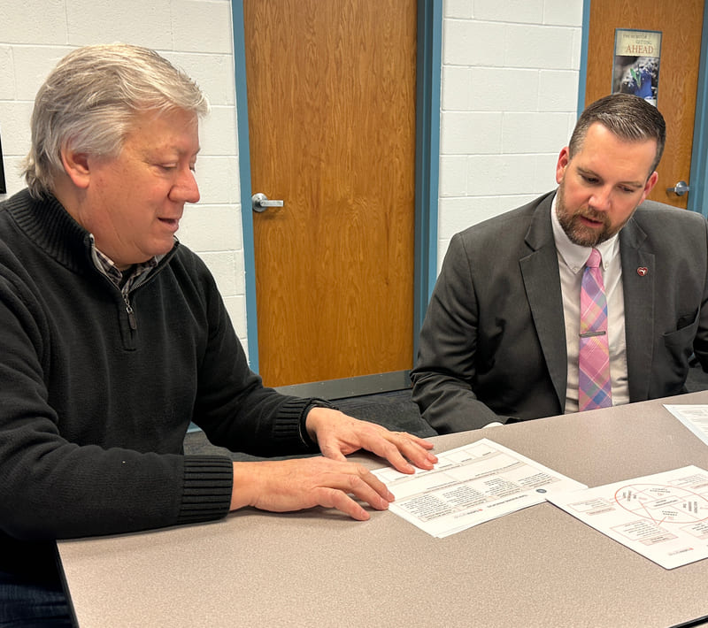 Mark sitting at a table with a customer, showing him charts and tables on paper placed in front of them